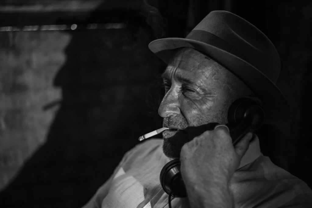 Man wearing hat is smoking a cigarette with old phone in hand. Film Noir Photography
